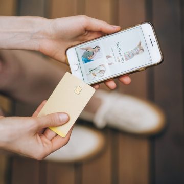 A shopper browsing clothes on a smartphone app while holding a gold credit card on a wooden bench.